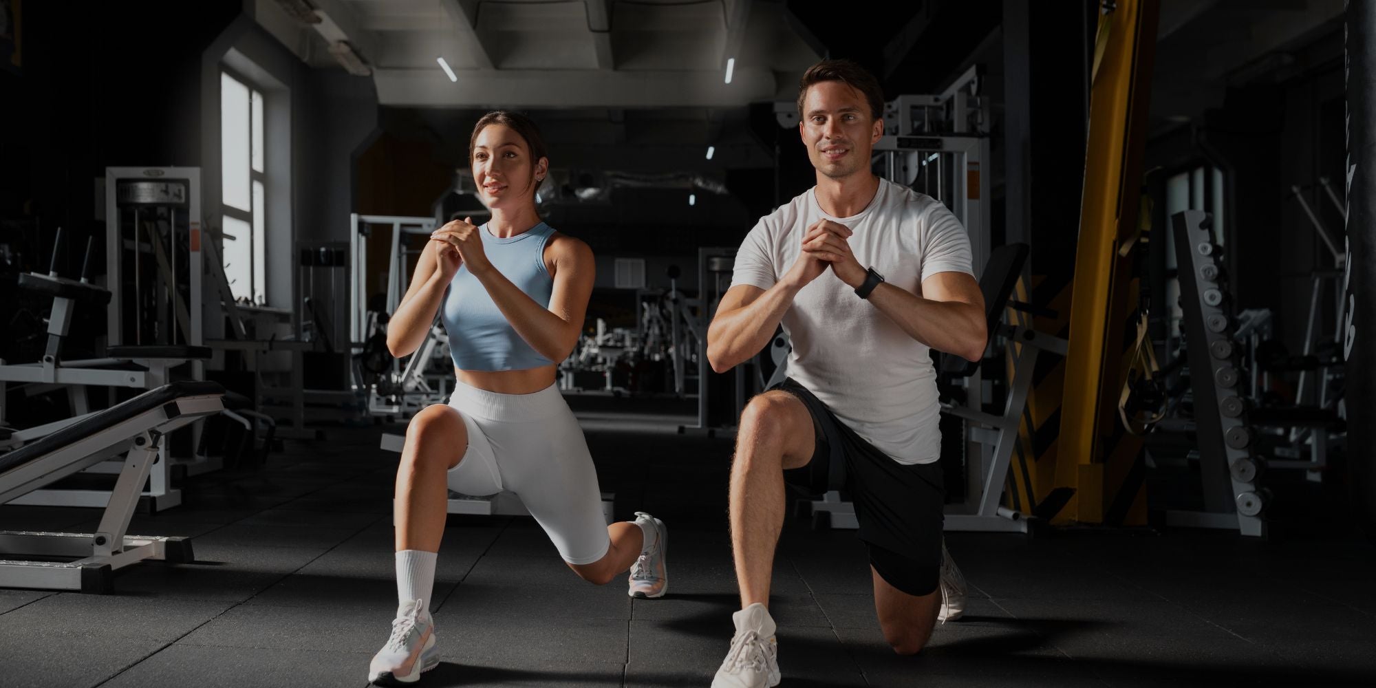 A man and a woman in athletic wear perform lunges in a dark gym setting with exercise equipment visible in the background.