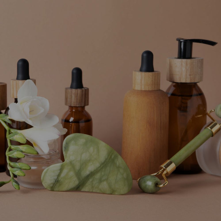 A set of natural skincare bottles and tools arranged on a warm beige background, including amber glass dropper bottles, pump bottles with light wood caps, a green jade roller, a green jade eye mask, and a white flower in a glass vase.