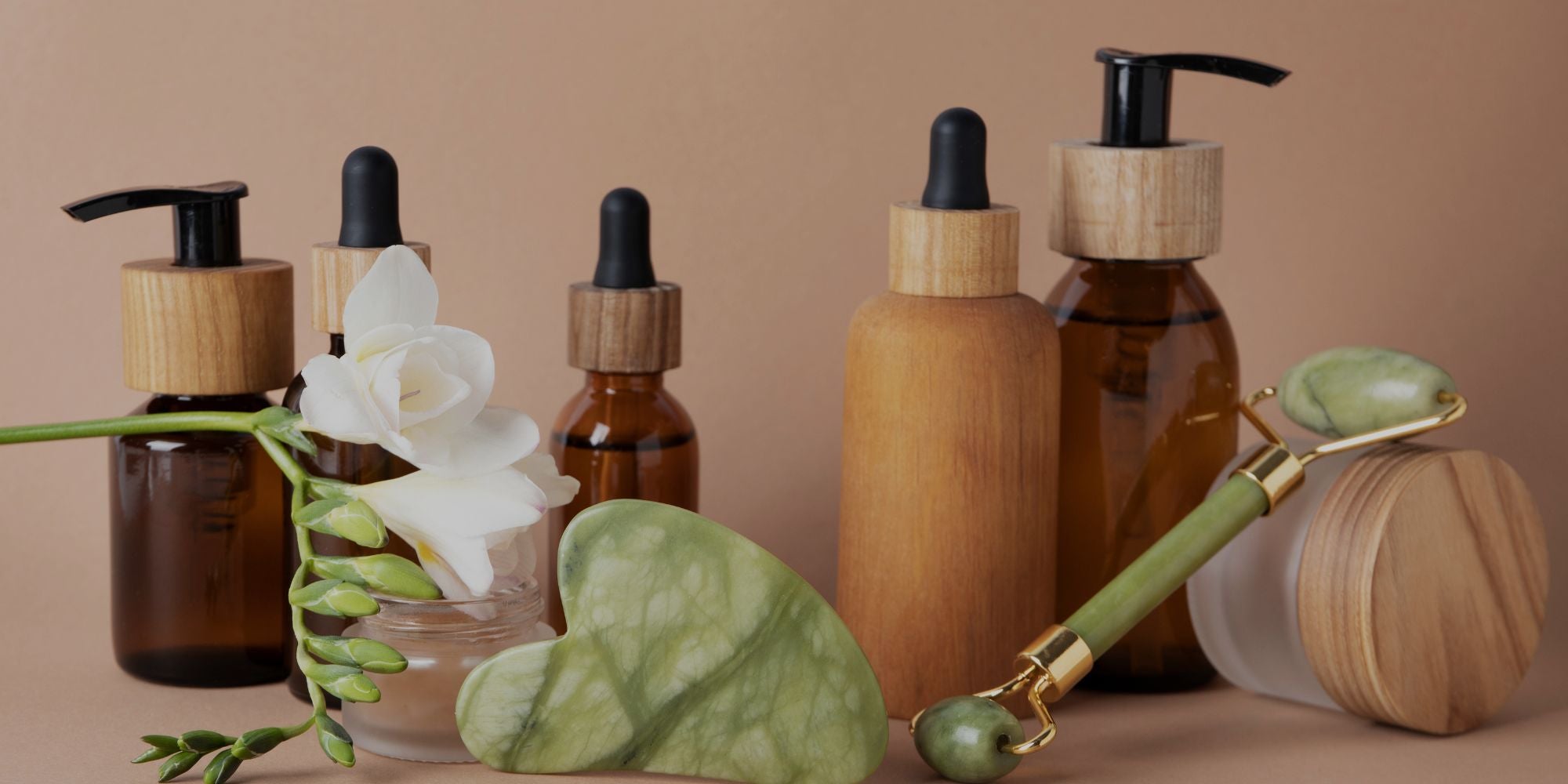 A set of natural skincare bottles and tools arranged on a warm beige background, including amber glass dropper bottles, pump bottles with light wood caps, a green jade roller, a green jade eye mask, and a white flower in a glass vase.