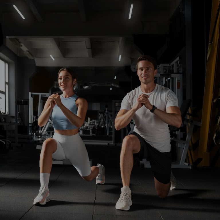 A man and a woman in athletic wear perform lunges in a dark gym setting with exercise equipment visible in the background.